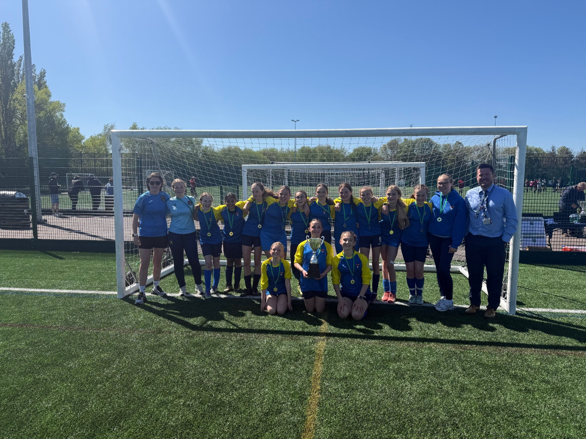 The Eckington School U12 Girls Football Team, Headteacher Mr Richard Cronin, coach Miss Robinson, and junior coaches Ella and Amelie smiling in a goal post with their medals and trophy
